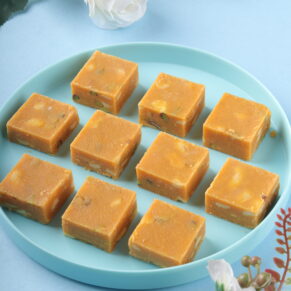 Square pieces of Besan Burfi made from gram flour, sugar, and ghee, garnished with chopped nuts, arranged on a decorative plate.