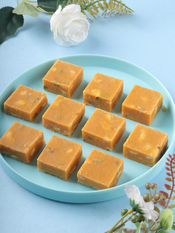 Square pieces of Besan Burfi made from gram flour, sugar, and ghee, garnished with chopped nuts, arranged on a decorative plate.