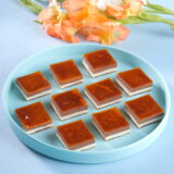 Square pieces of mango chocolate delight burfi, layered with mango and chocolate flavors, decorated with chopped nuts, arranged on a decorative plate.